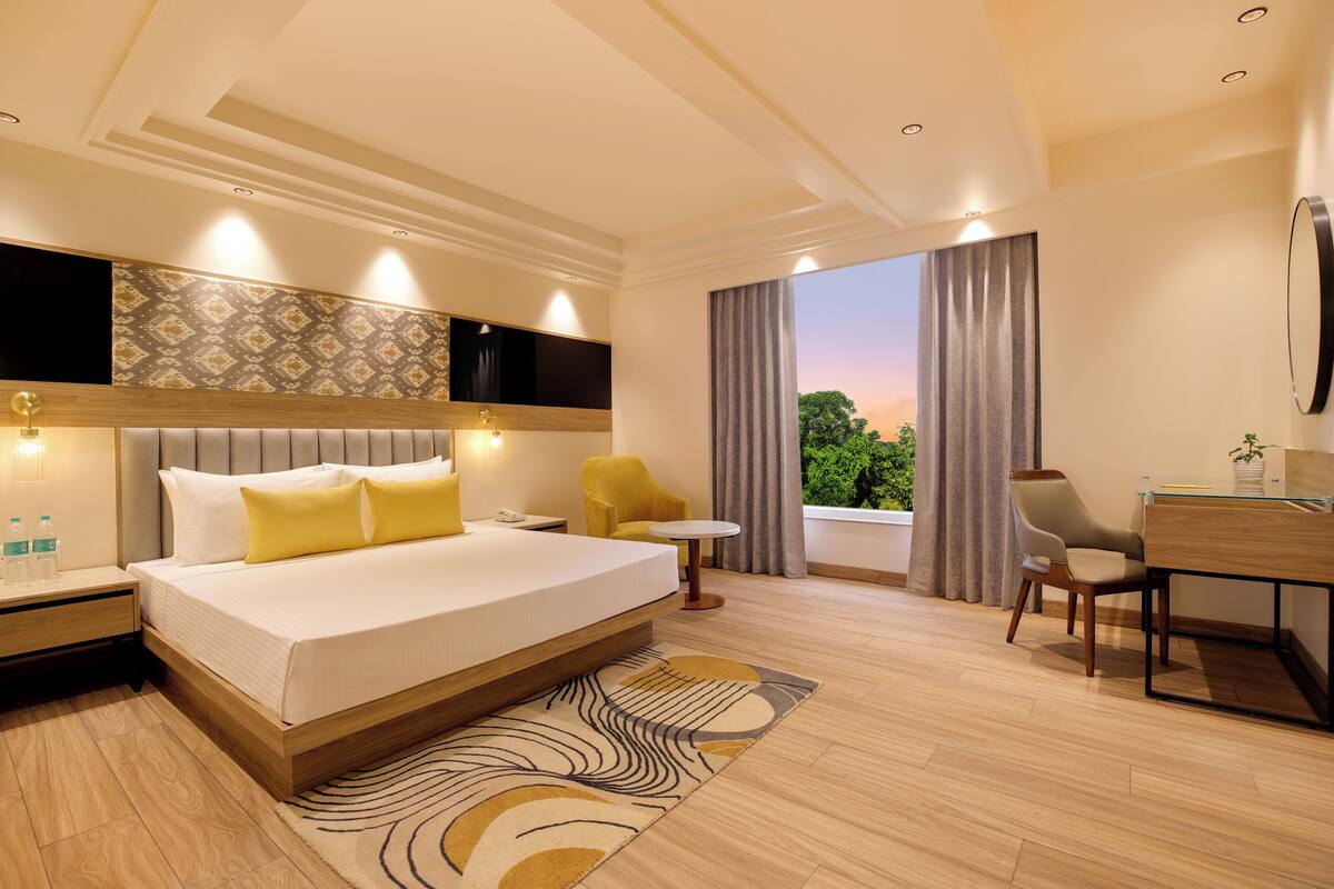 A modern hotel room with a large bed, a desk, a chair, a small table, and a view of greenery.