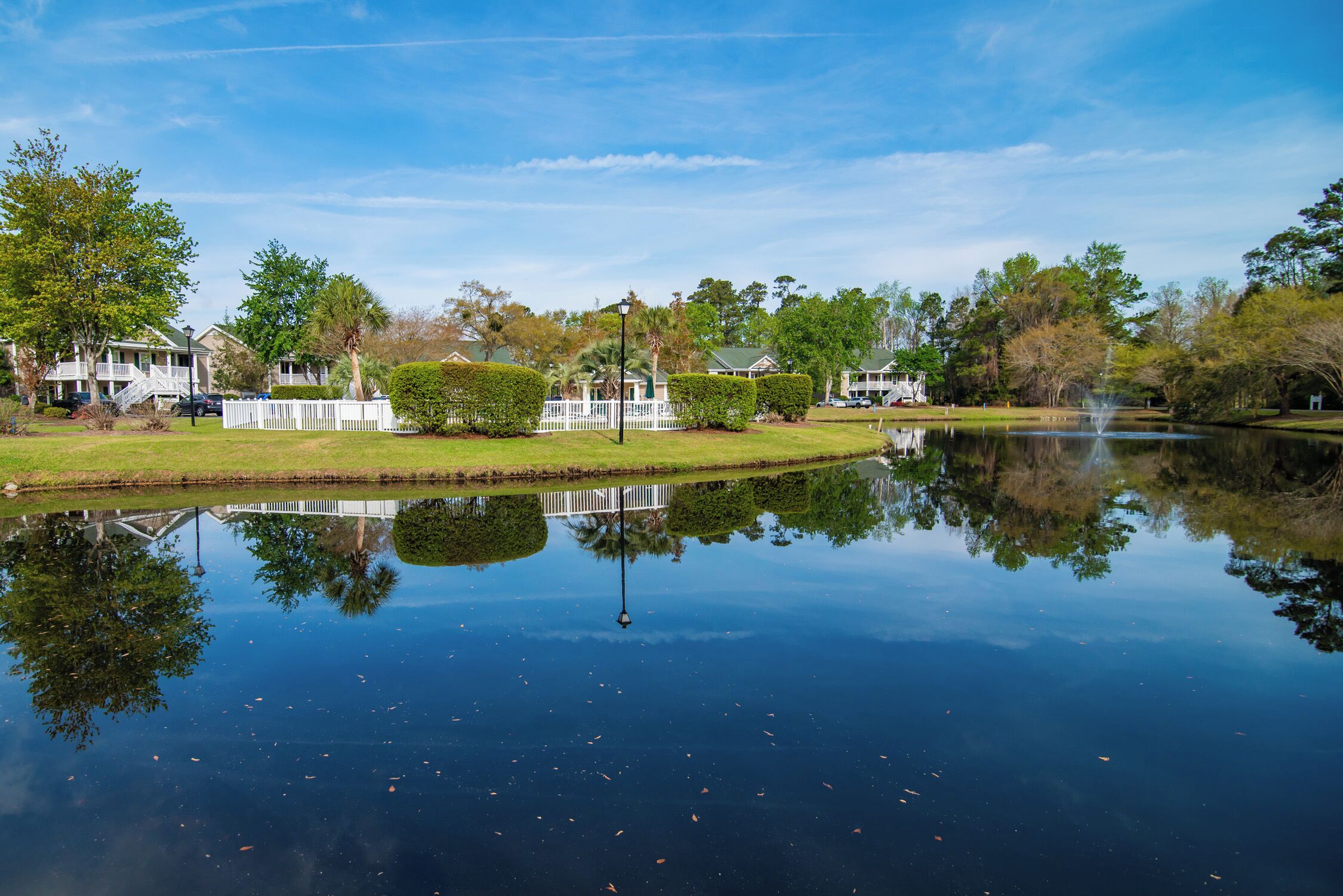A calm lake with a reflection of trees and buildings, surrounded by well-maintained lawns and a white fence.