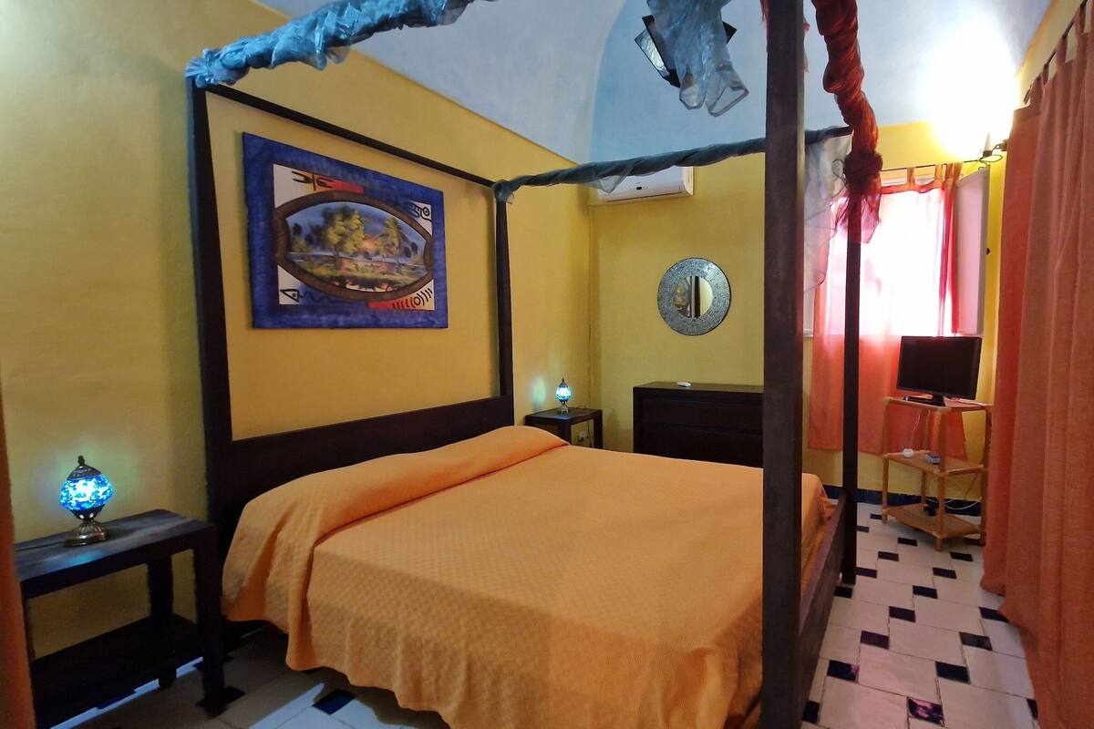Ein Zimmer mit einem Himmelbett, einem Bild an der Wand, einem kleinen Tisch mit Lampe, einem Spiegel und einem Fernseher auf einem Standfuß.