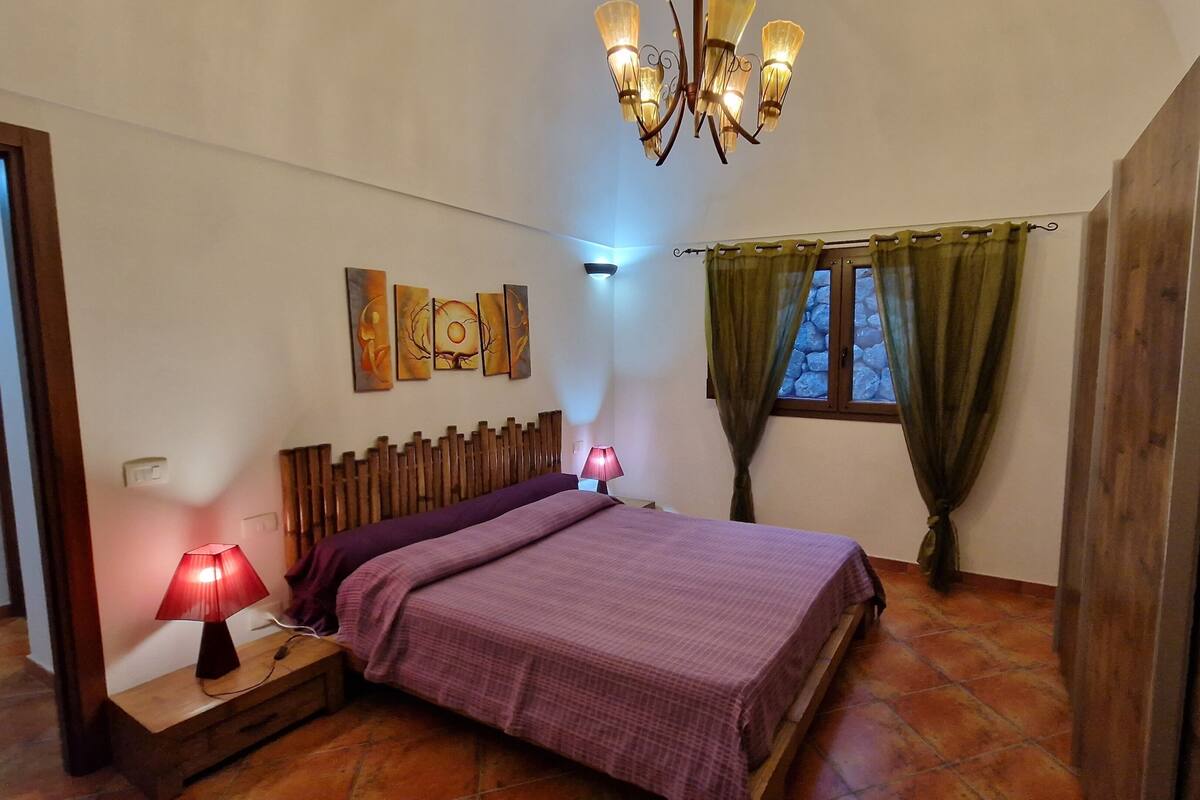 Ein Schlafzimmer mit einem Bett, zwei Nachttischlampen, einer Deckenleuchte, einem Fenster mit Vorhängen und einer Wand mit Kunstwerken.