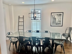 A dining area with a glass table and black chairs, a ladder, a framed cityscape picture, and a window with curtains.