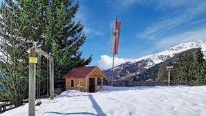 A wooden cabin with a red roof, surrounded by snow-covered ground and trees, with a flag in the background.
