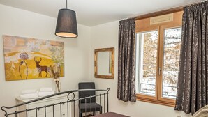 Une chambre à coucher comprenant un lit, un bureau, une chaise, un luminaire suspendu, une fenêtre avec des rideaux et un tableau accroché au mur.
