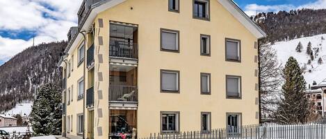 Un immeuble à plusieurs étages doté de balcons, un sol recouvert de neige et un paysage montagneux en arrière-plan.