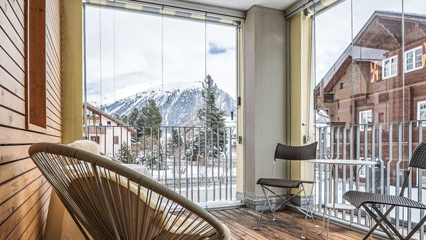 Un balcon doté d’un plancher en bois, d’une chaise en osier, d’une table en métal et offrant une vue sur des montagnes enneigées.