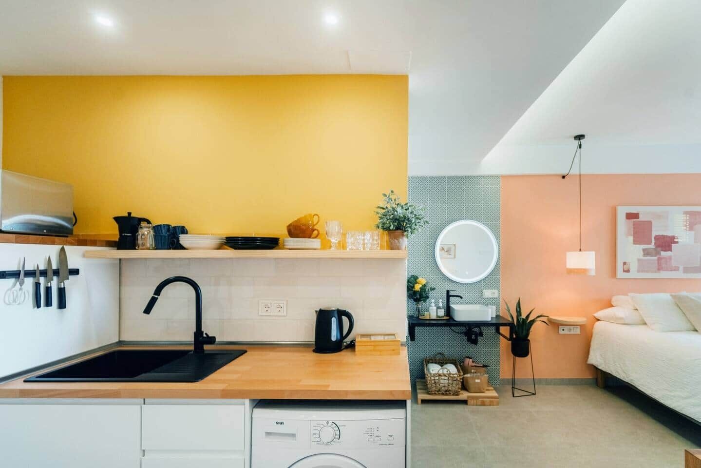 Een compacte keuken met een spoelbak, fornuis en wasmachine, een klein zite hoek met een tafel en stoelen, en een slaapkamer met een bed en nachtkastje.