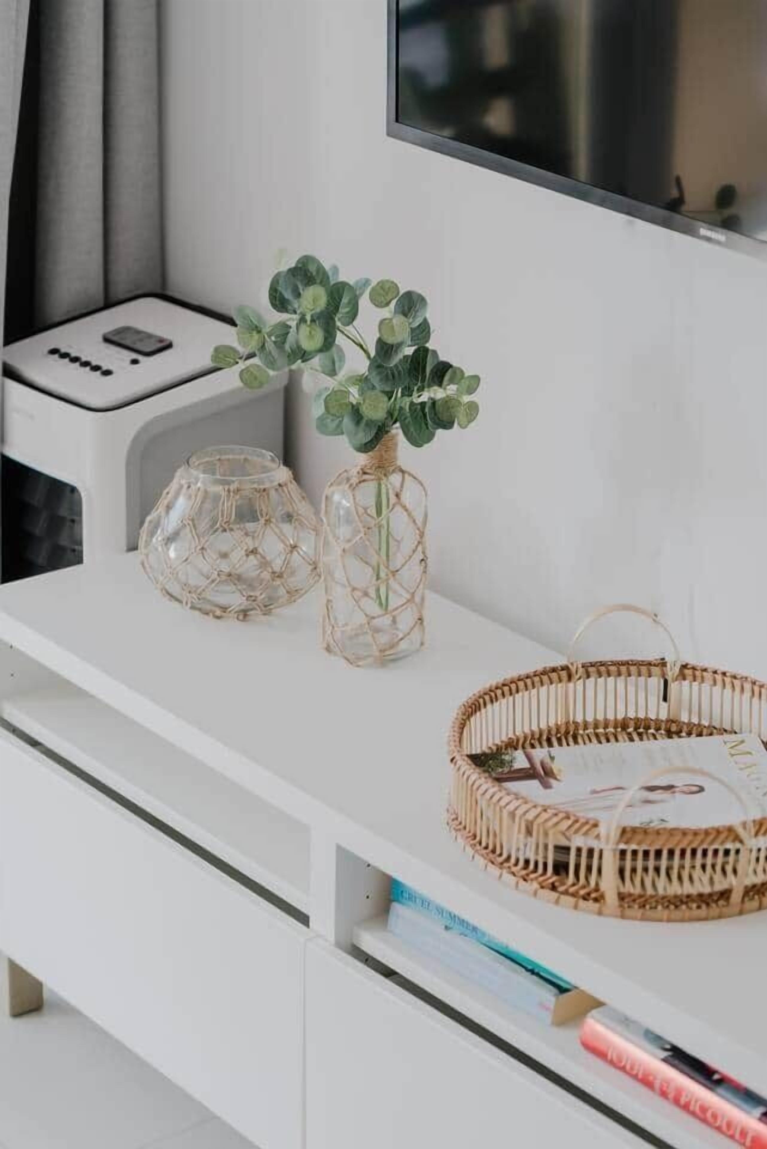 Et hvidt skrivebord med en flettet kurv, to glasvaser med planter og et tv ophængt på væggen.