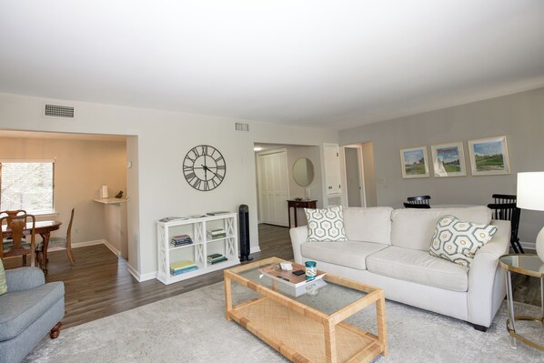 A living room with a white sofa, a coffee table, a bookshelf, and a dining area with chairs and a table.