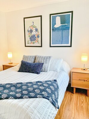 Ein ordentlich gemachtes Bett mit einer blau-weißen Bettdecke, zwei Kissen und einer blauen Tagesdecke. Zwei Nachttische mit Lampen und zwei gerahmte Kunstwerke an der Wand sind ebenfalls vorhanden.