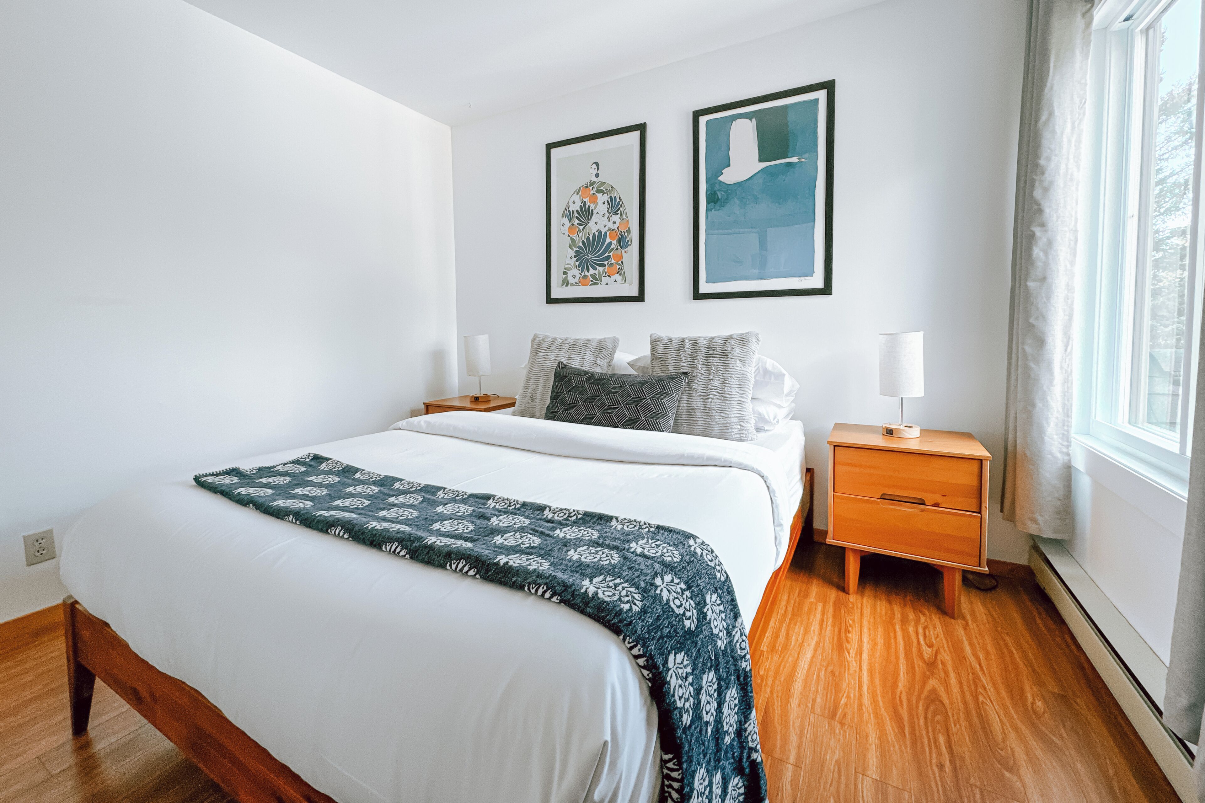 A bedroom with a bed, bedside tables, a nightstand, a window, and two framed artworks on the wall.