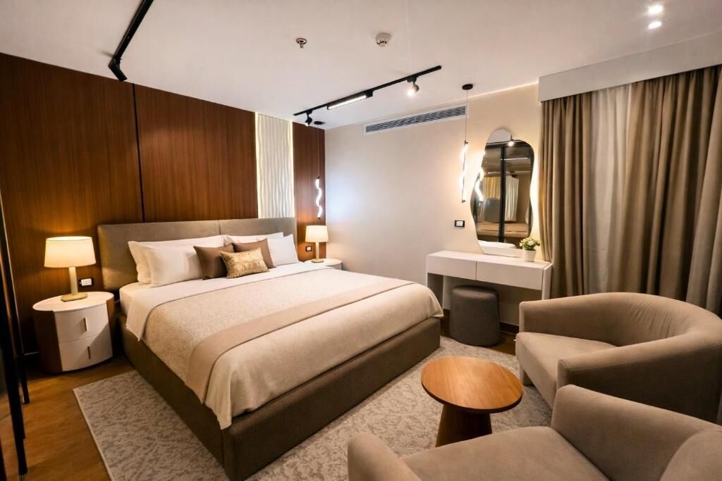 A modern hotel room with a large bed, bedside tables, a vanity, and a seating area.