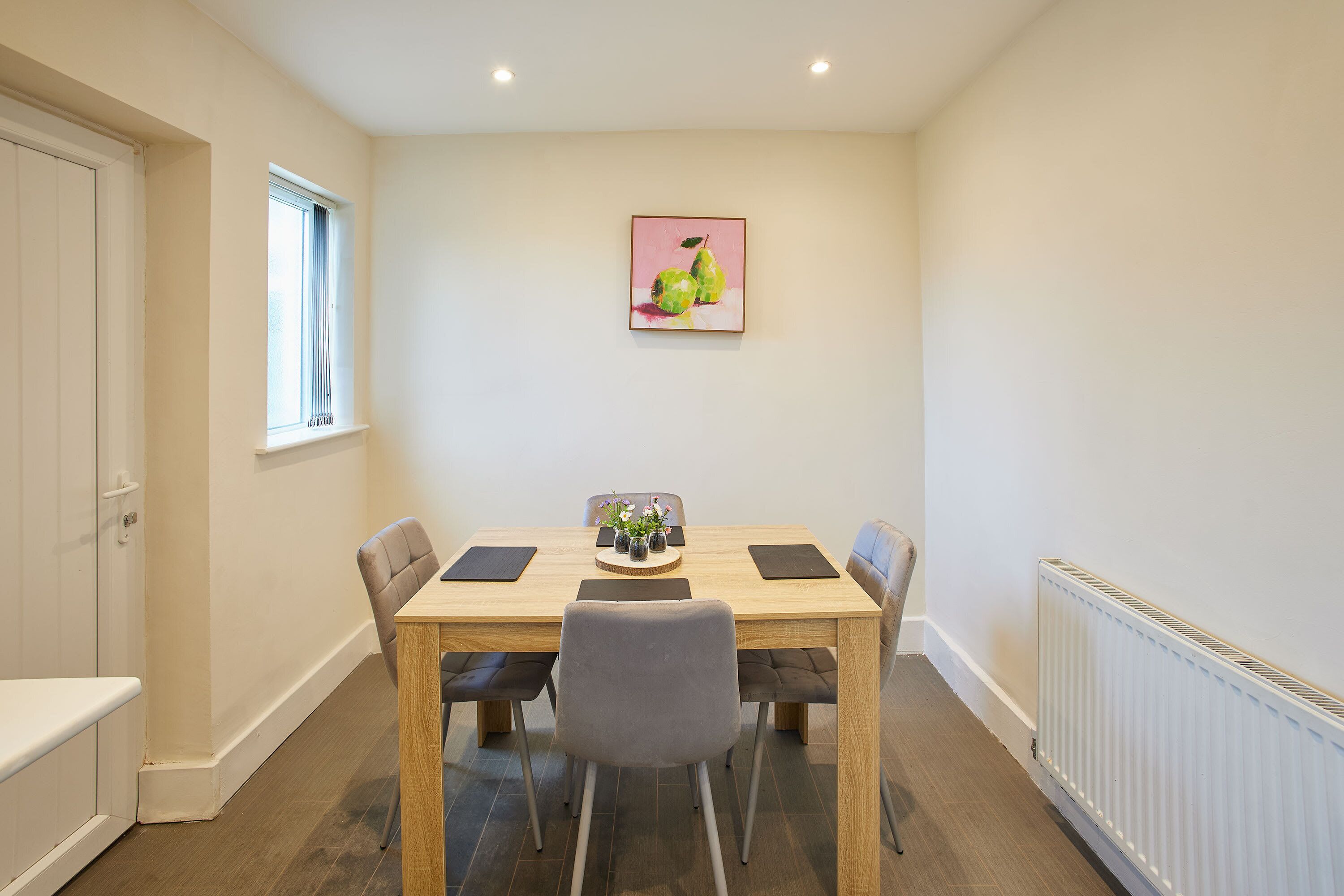 A dining area with a wooden table, four chairs, a vase with flowers, and a painting on the wall.