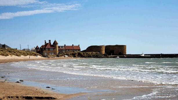 Un paisaje costero con una playa arenosa, un edificio grande con torreón y una estructura que parece una fortaleza.
