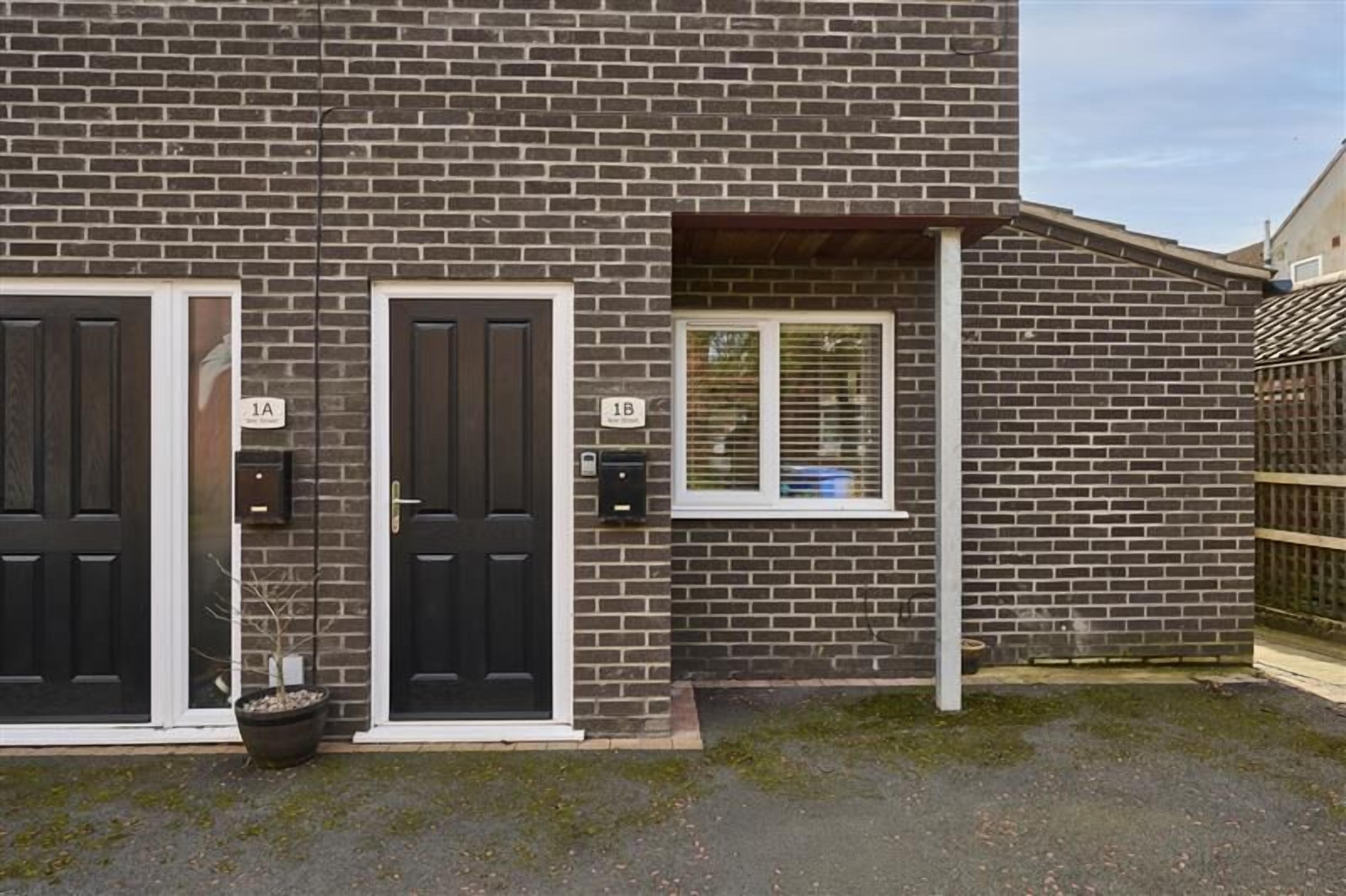 Un bâtiment en briques de deux étages, doté de deux portes noires portant les inscriptions « 1A » et « 1B », d’une petite fenêtre avec des stores et d’un pot de fleurs placé devant la porte « 1A ».