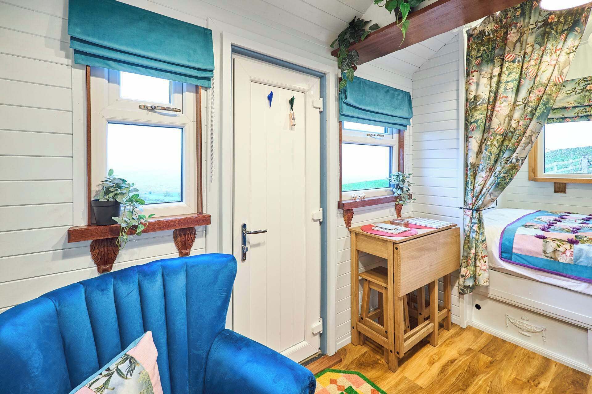 A room with a blue chair, a wooden table, and a bed with floral curtains.