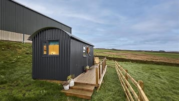 A small, dark-colored cabin with a wooden deck, situated on a grassy field with a large building in the background.