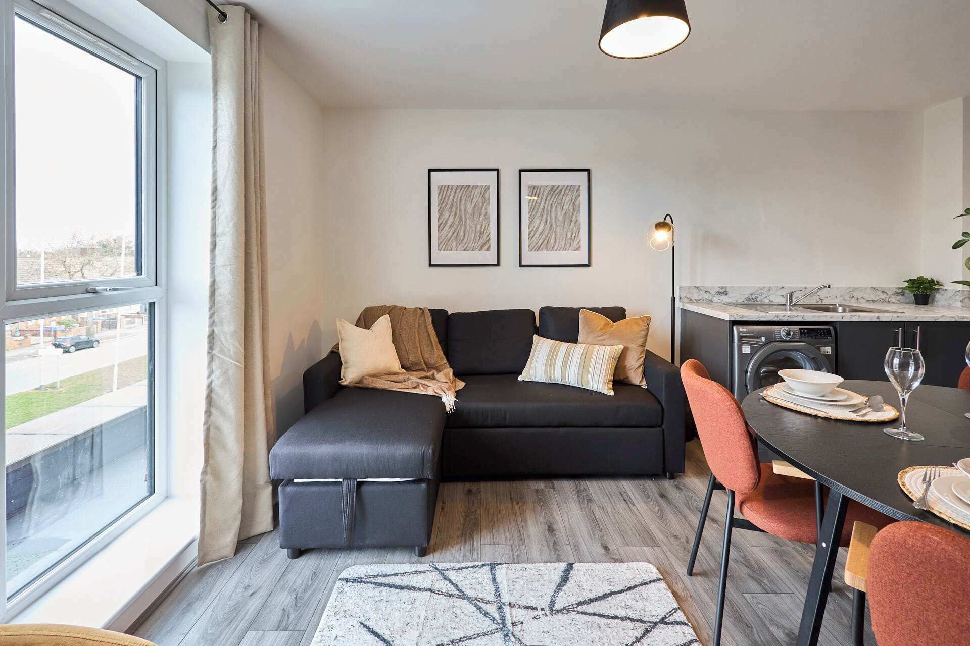 Un espace de vie moderne comprenant un canapé, une table à manger avec des chaises, un lave-linge et deux œuvres d’art encadrées accrochées au mur.
