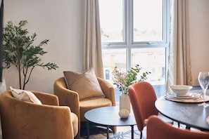 Ein gemütliches Zimmer mit einer Couch, Sesseln, einem kleinen Tisch und einem Fenster mit Blick auf Gebäude.