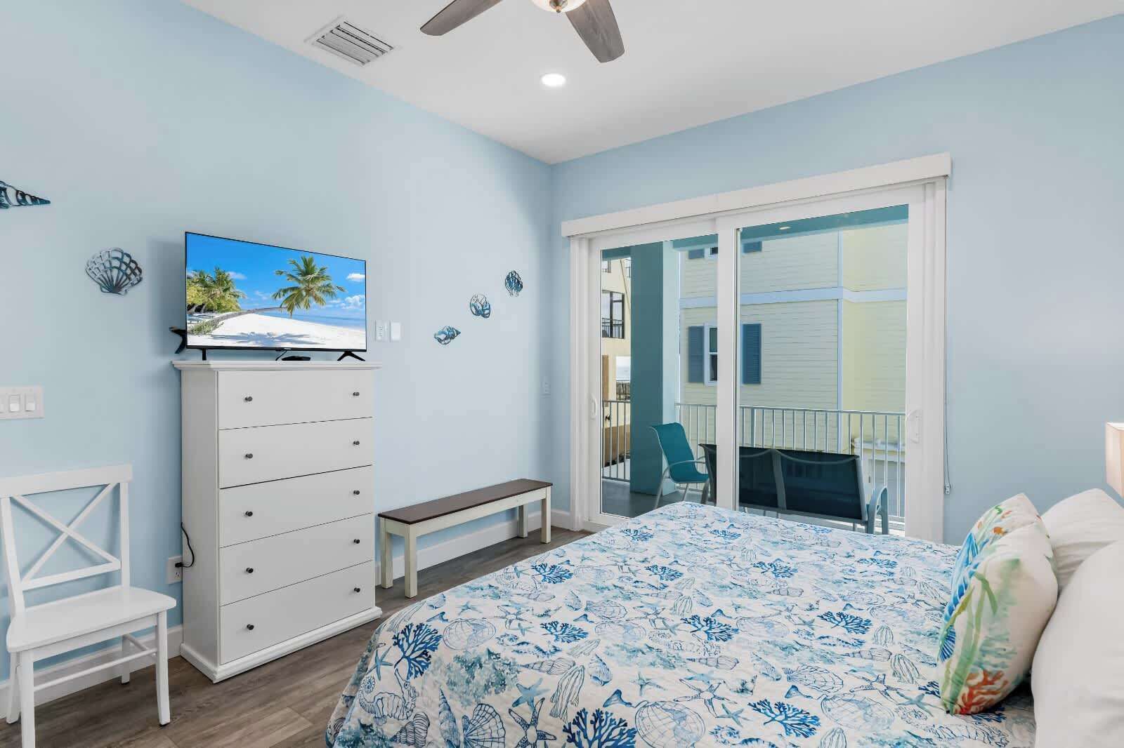 A bedroom with a bed, a dresser, a bench, a TV, and a balcony with a chair.