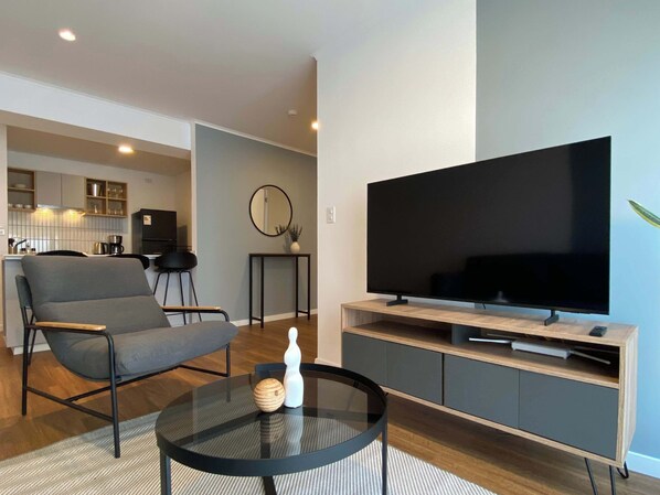 Een moderne woonkamer met een flatscreen-tv, een grijze bank, een glazen salontafel, en een keuken op de achtergrond.