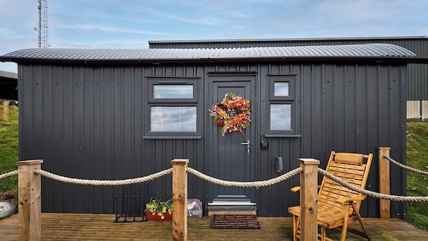 A dark wooden building with a metal roof, a wreath on the door, a wooden chair, and a rope barrier. - Host Stay Curlews Rest (Preston)