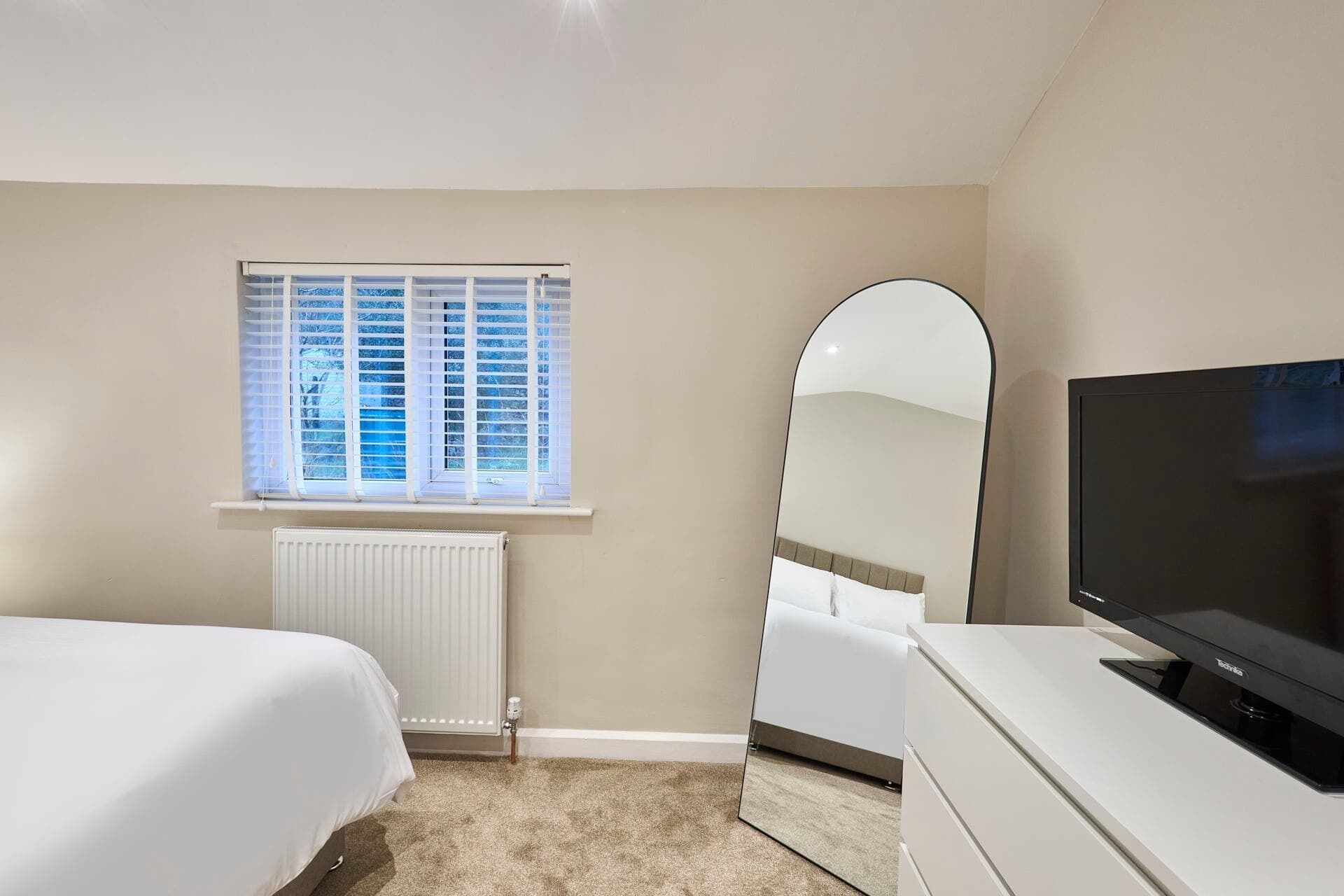 Een slaapkamer met een bed, een dressoir, een televisie, een spiegel en een raam met jaloezieën.