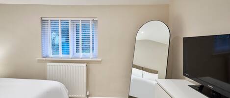 Een slaapkamer met een bed, een dressoir, een televisie, een spiegel en een raam met jaloezieën.