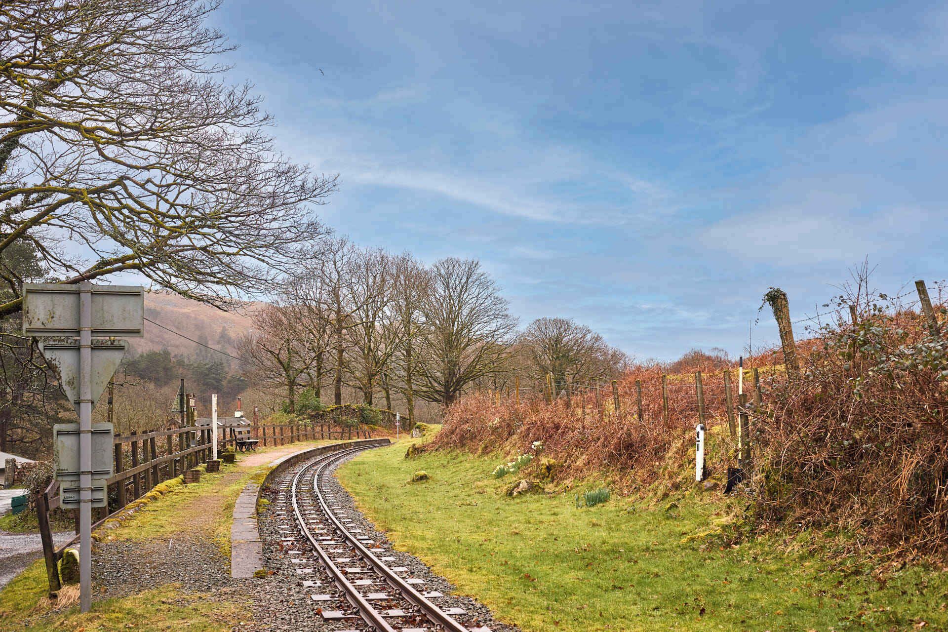 Vías de tren serpenteando a través de un paisaje rural con una cerca de madera y árboles desnudados.