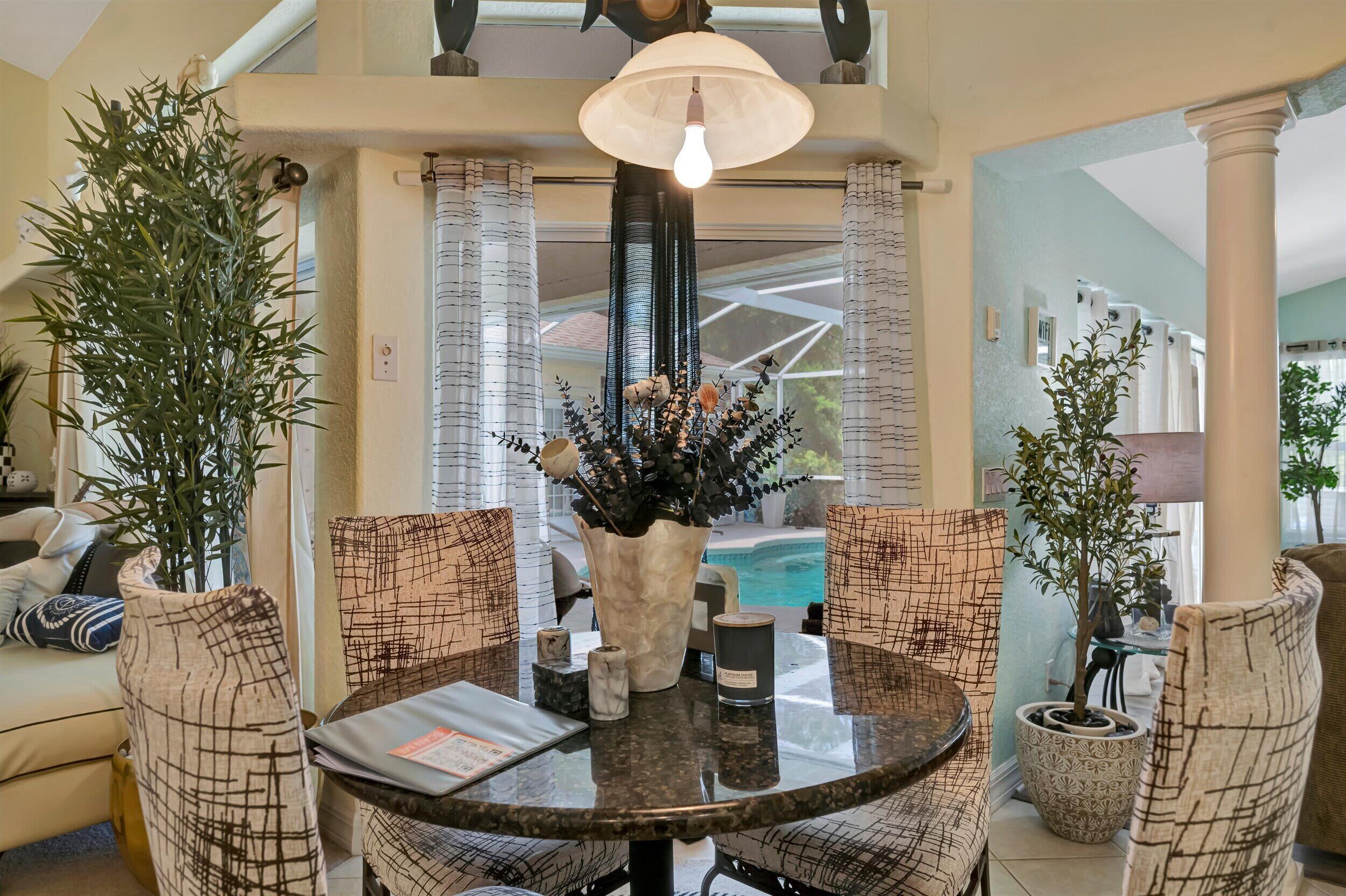 A dining area with a round table, chairs, a vase with flowers, and a view of a swimming pool.