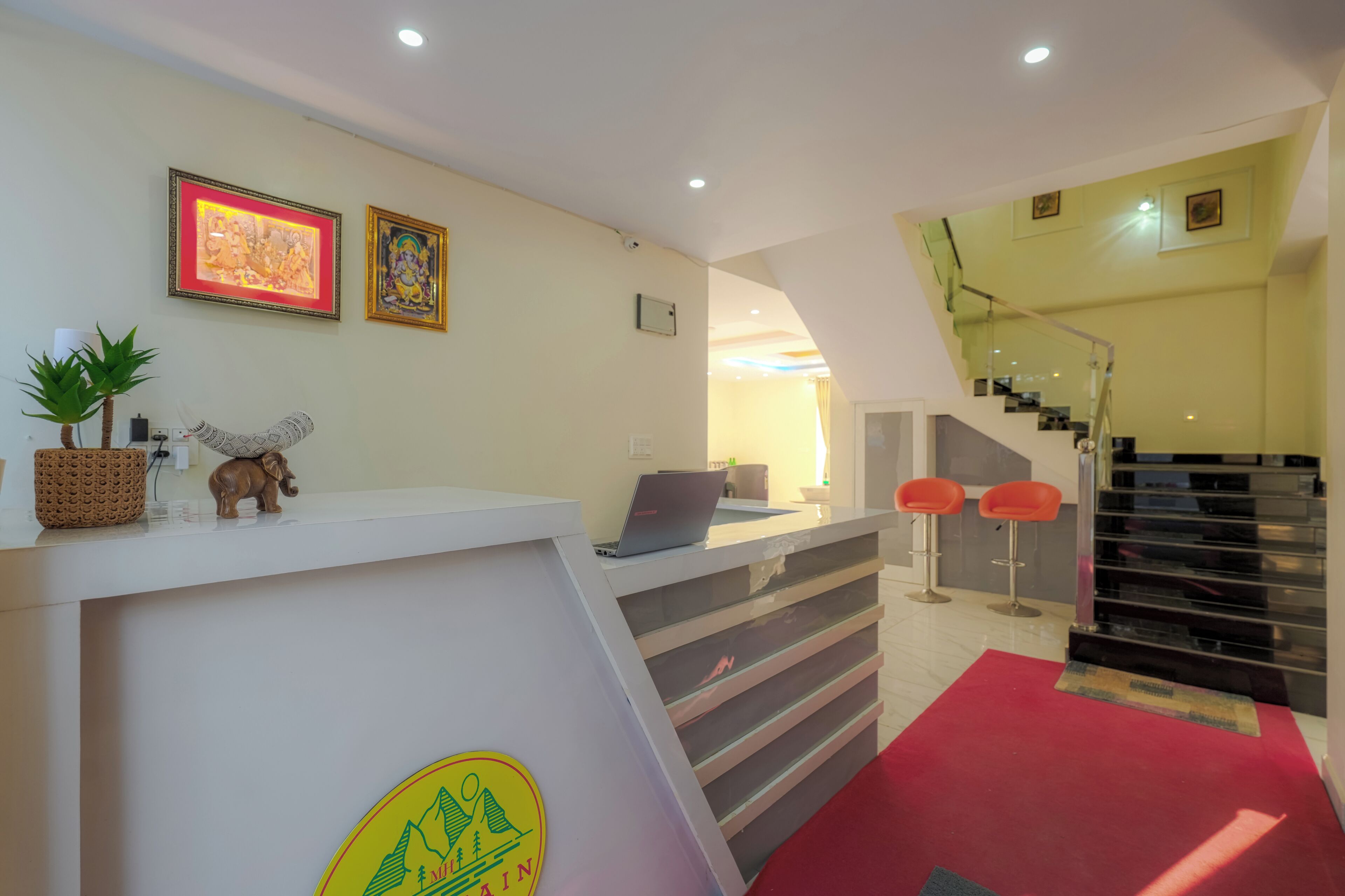 A reception desk with a laptop, framed artwork, and a logo. A staircase with a red carpet leads to a second floor.