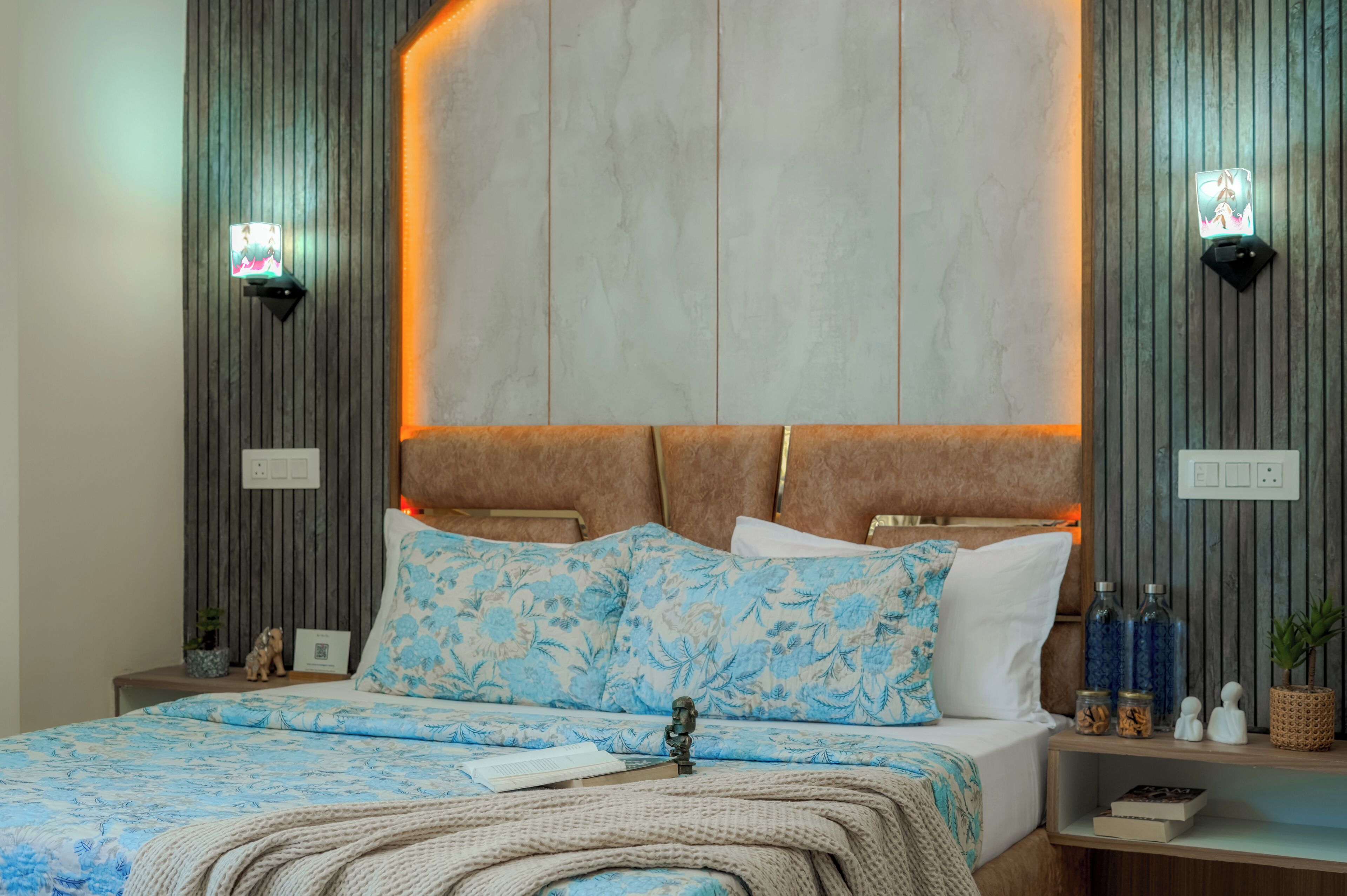Un dormitorio moderno con una cama grande, cabecera con iluminación integrada, mesita de noche con una planta y mesita auxiliar con adornos.