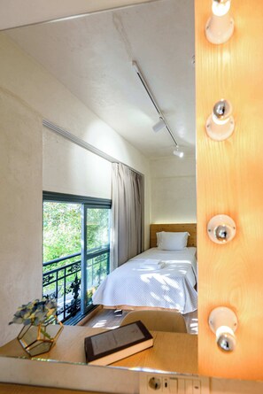 Ein Hotelzimmer mit Bett, Schreibtisch mit Lampe, Vorhängen an einem Fenster und Blick ins Grüne.