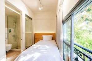 Ein Schlafzimmer mit Bett, Fenster mit Blick auf Grünflächen, einer Duschkabine und einem Holzkopfende.