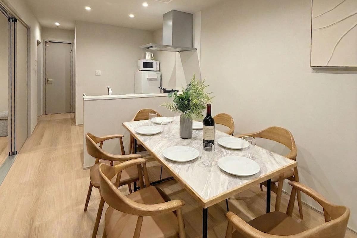 A dining area with a table set for six, wooden chairs, a kitchen in the background, and a white wall with a simple artwork.