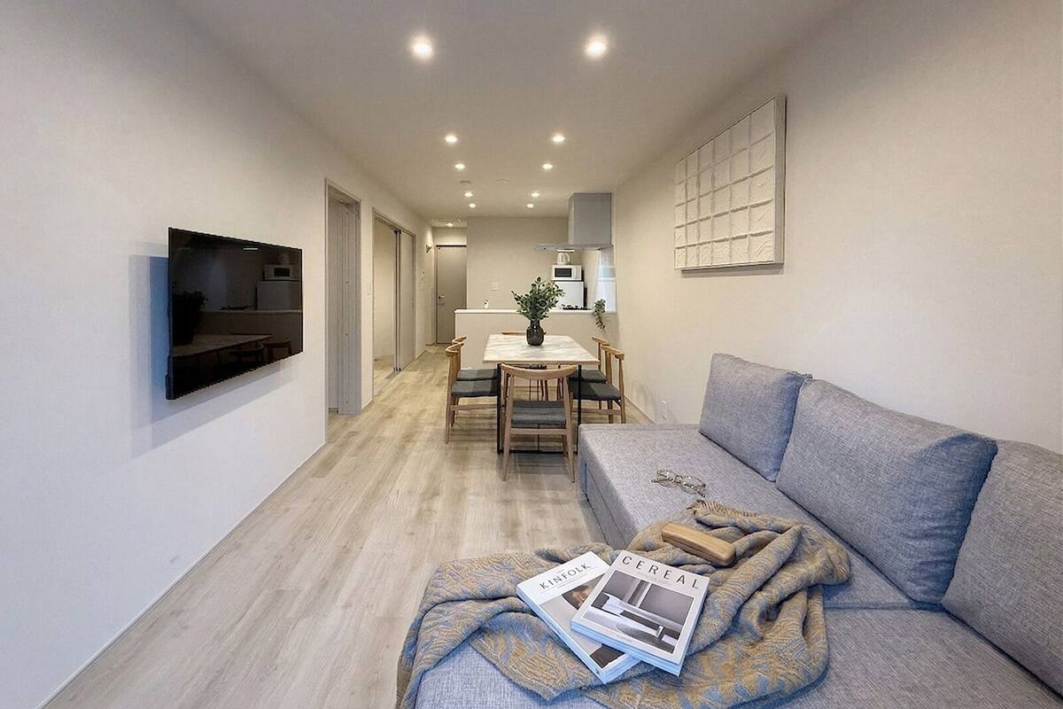 A modern living room with a sofa, a dining area with a table and chairs, a kitchen in the background, and a flat-screen TV mounted on the wall.