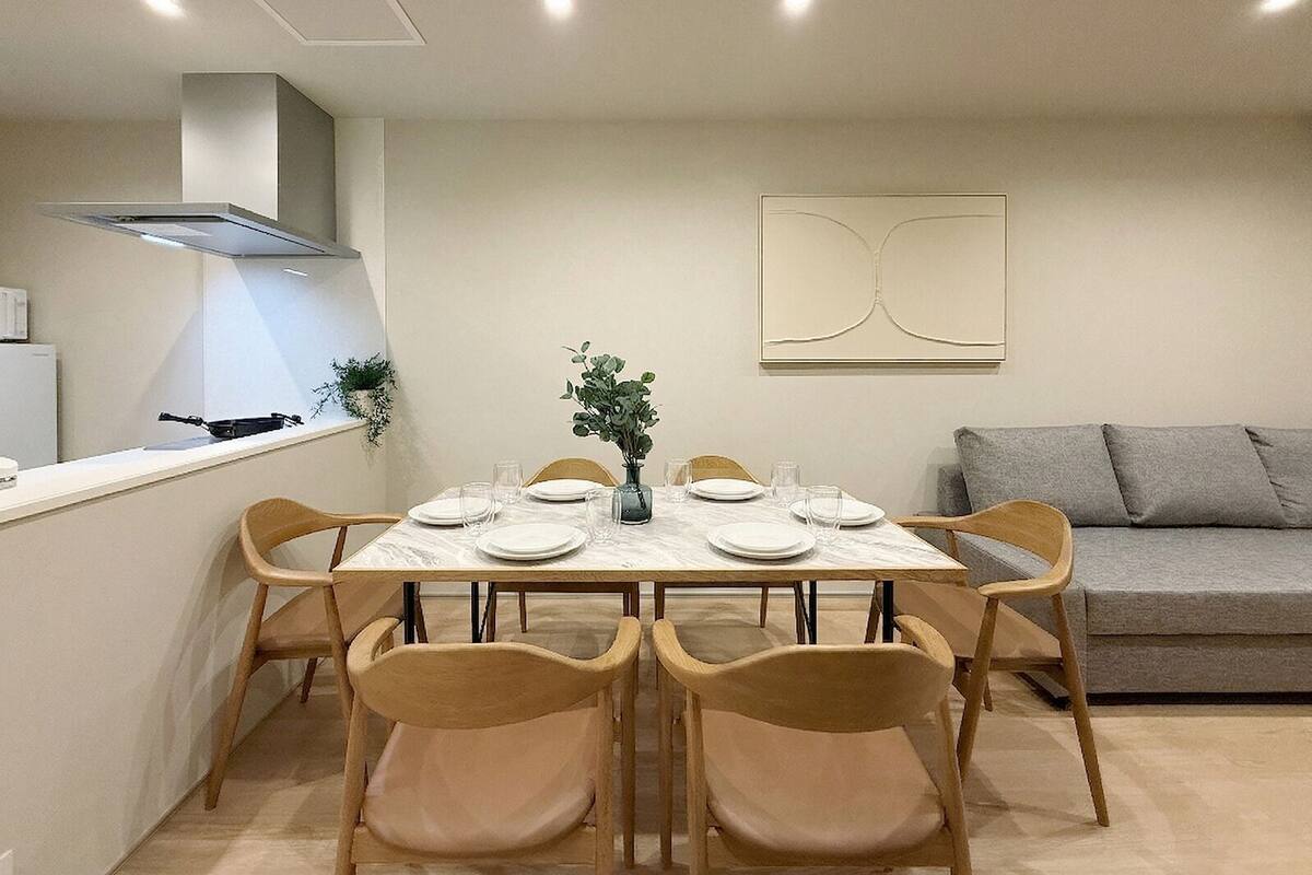 A dining area with a marble table, six wooden chairs, a vase with greenery, and a modern kitchen in the background.