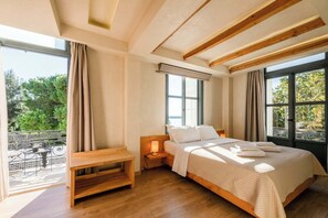 A bedroom with a wooden bed, a nightstand with a lamp, a bench, and large windows offering a view of greenery. - Panellinion Beachfront Getaway - Sea Sunny Gem (Kalamata)