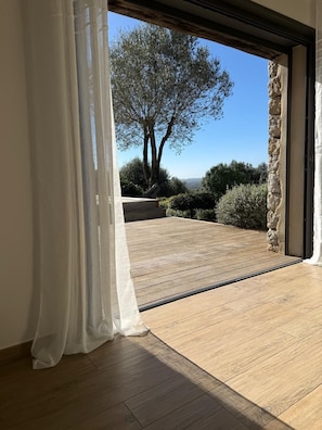 A room with a wooden floor, a stone wall, and a view of a tree and landscape. - Quiet Lucia sheepfold, private heated pool (Figari)