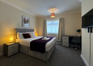 Quarto de hotel com cama, mesa de cabeceira, escrivaninha, cadeira e televisão.