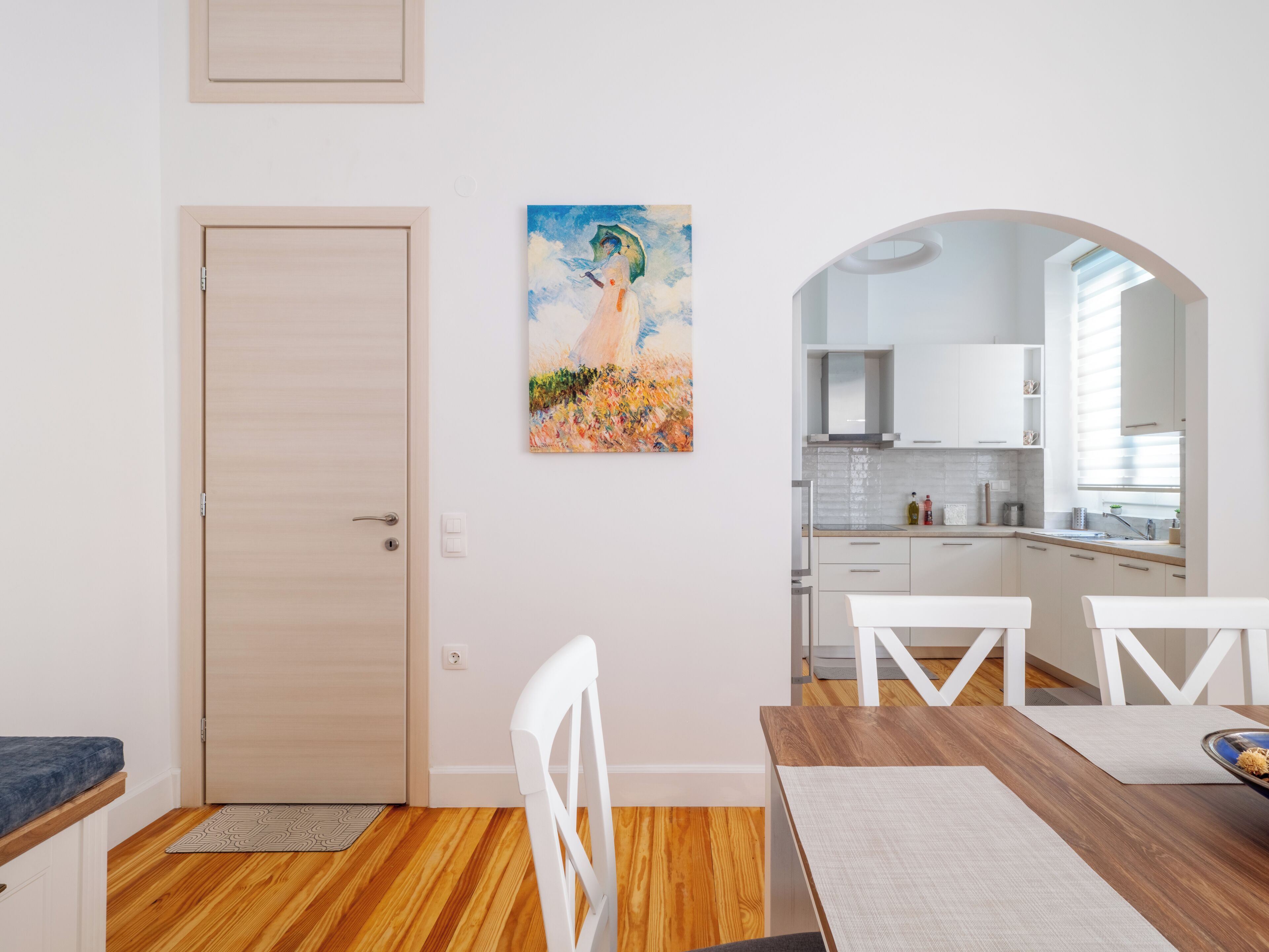 A modern kitchen with white cabinets, a wooden dining table, and a painting on the wall.