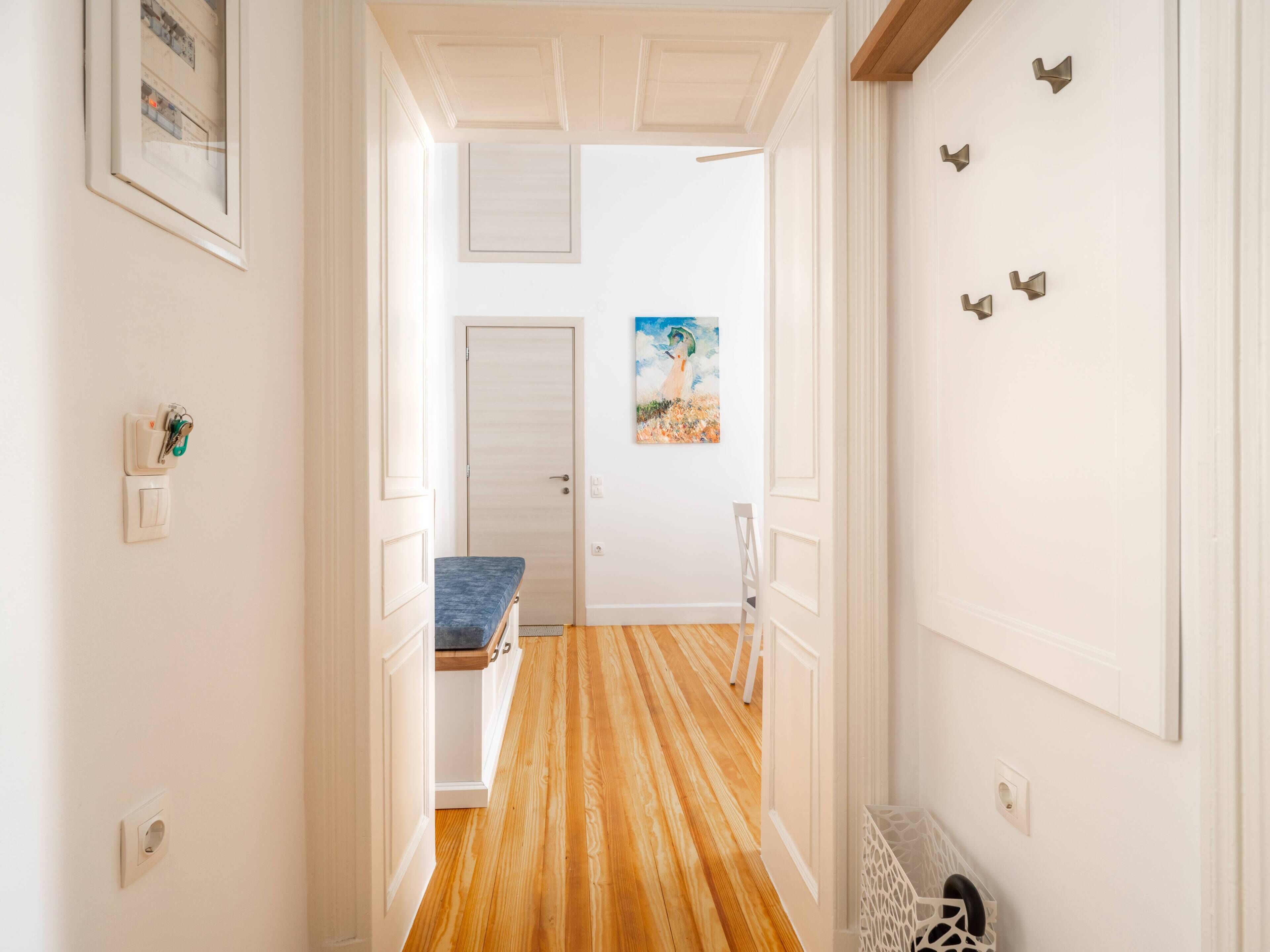 A hallway with a wooden floor, white walls, a door, a bench, and a painting on the wall.