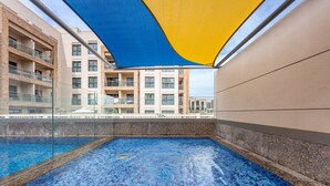 Ein Schwimmbecken mit blauem Mosaikboden, einer Glasufernung und Blick auf ein mehrstöckiges Gebäude mit Balkonen.