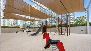 A playground with a slide and swings, set against a backdrop of modern buildings. - Free Housekeeping Visits During Your Stay - Stayshort - Relaxing 1 Bed That Sleeps 4 With Amazing Views From Balcony (Dubai)
