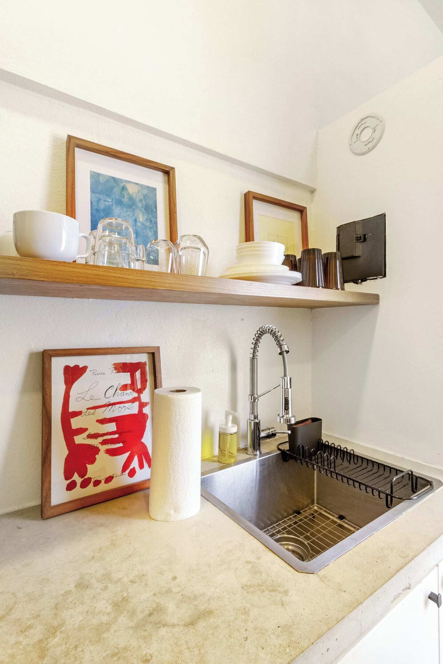 A kitchen with a stainless steel sink, a wooden shelf with dishes, a framed artwork, and a roll of paper towels.