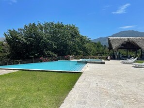 Piscina com espreguiçadeiras, uma estrutura com telhado de palha e montanhas ao fundo.