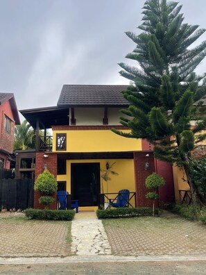 Uma casa de dois andares com fachada amarela e vermelha, um alpendre coberto com cadeiras azuis e uma grande árvore no jardim da frente.