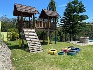 Une structure de jeu en bois, dotée d’un toboggan et d’un mur d’escalade, entourée d’herbe verte et d’arbres.
