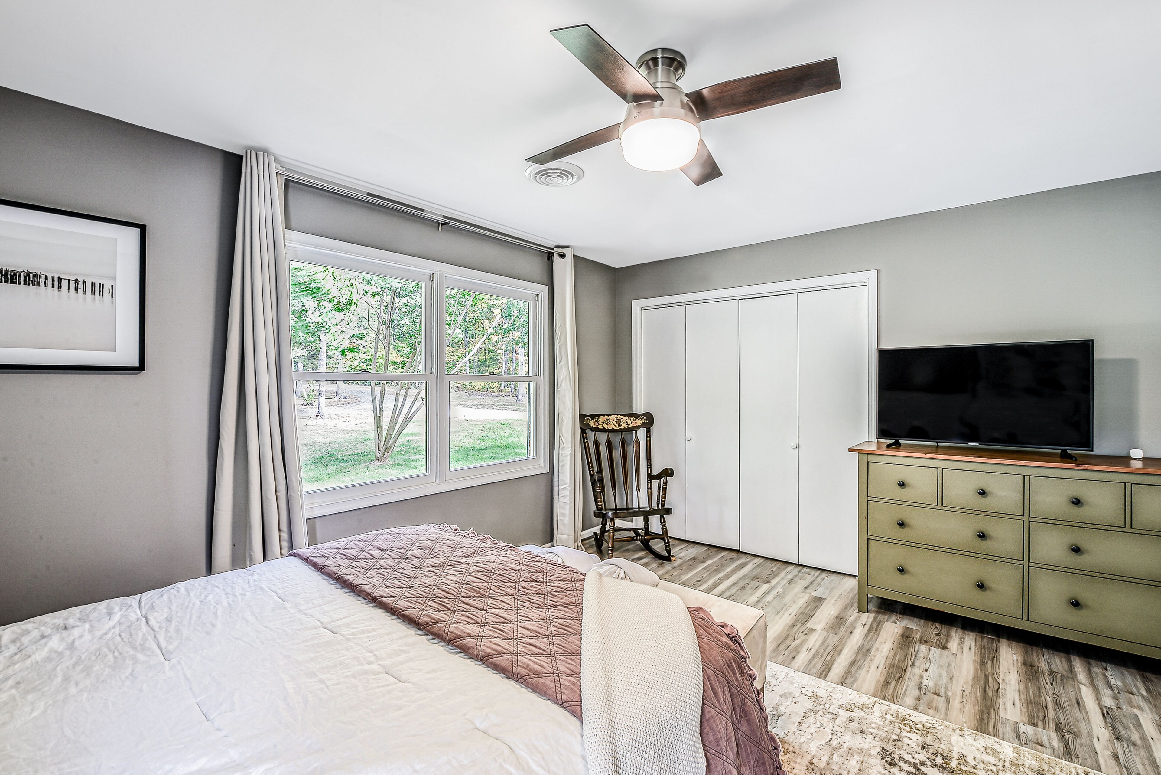 Un dormitorio con cama, ventilador de techo, televisor sobre una cómoda y ventana con cortinas.
