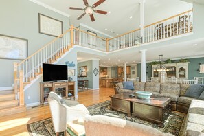 A spacious living room with a high ceiling, a staircase, and a kitchen in the background.