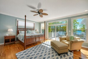 Un dormitorio con cama con dosel, ventilador de techo, ventana grande con vista a árboles y balcón con mobiliario exterior.